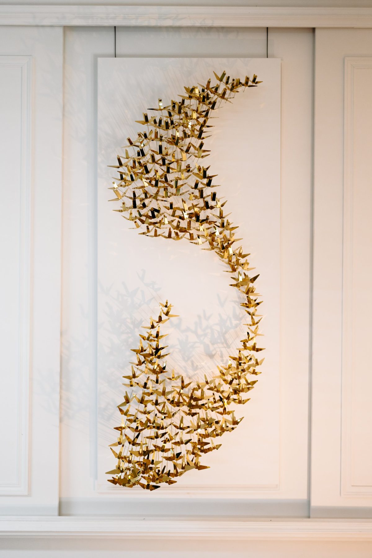 Polished brass Murmuration: a vertical, wave-shaped flock on a white panel.